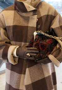 Bruin- en beige geruit wollen jas met een hoge kraag, houdt een zwarte clutch met bloemenpatroon en gouden ketting vast. Bruine leren handschoenen gedragen.