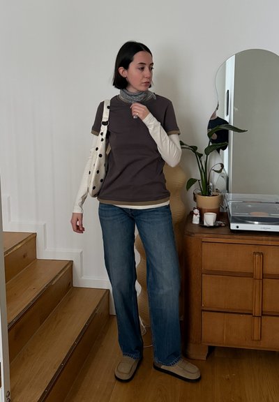 Mujer con cabello corto y oscuro, vestida con capas de prendas, vaqueros y zapatos beige, de pie junto a una cómoda de madera con una planta, girando ligeramente hacia la izquierda.