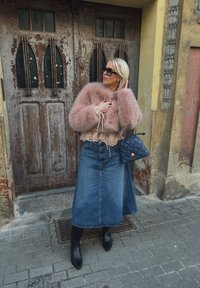 Femeie cu jachetă roz din blană, fustă lungă din denim, cizme negre, ochelari de soare și geantă albastră, stând pe trotuar în fața unor uși vechi din lemn.