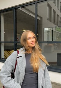 Cappotto di lana grigio con una vestibilità morbida, indossato sopra un top scuro. Il cappotto presenta grandi bottoni neri e un colletto ampio, abbinato a una borsa di colore rosso vino.