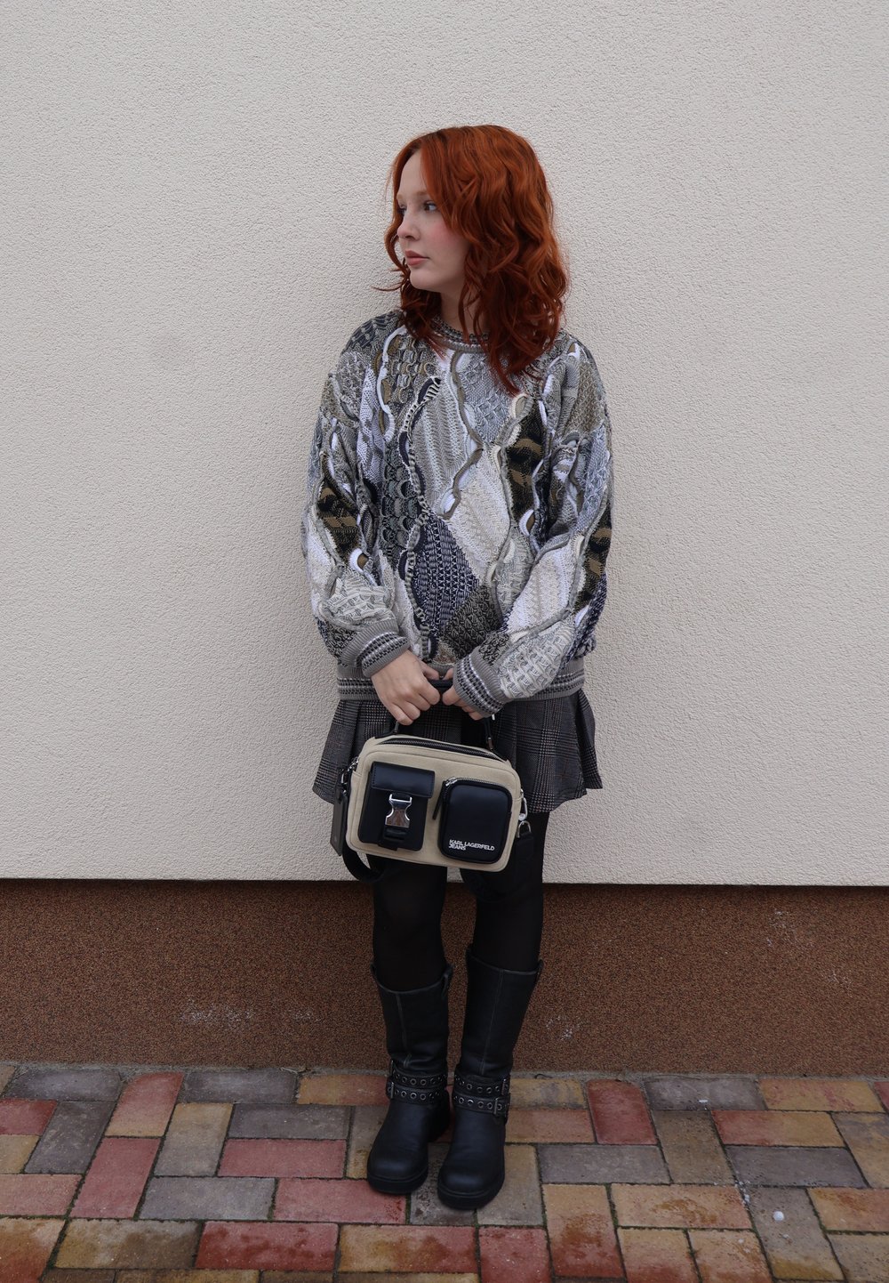 Femme aux cheveux roux bouclés portant un pull à motifs, une jupe à carreaux, des collants noirs, des bottes, tenant un sac à main beige et noir contre un mur.