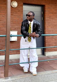 Casaco de couro preto sobre uma camisa branca, gravata com rosto sorridente amarelo, calças largas brancas, óculos de sol e ténis brancos. Fundo de tijolos.