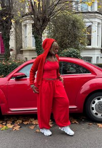 Sarkans adīts kapuču džemperis, sarkans bezroku tops un vaļīgas sarkanas sporta bikses. Balti sporta apavi ar teksturētām zolēm stāv blakus sarkanai automašīnai, rudens lapas izkaisītas.