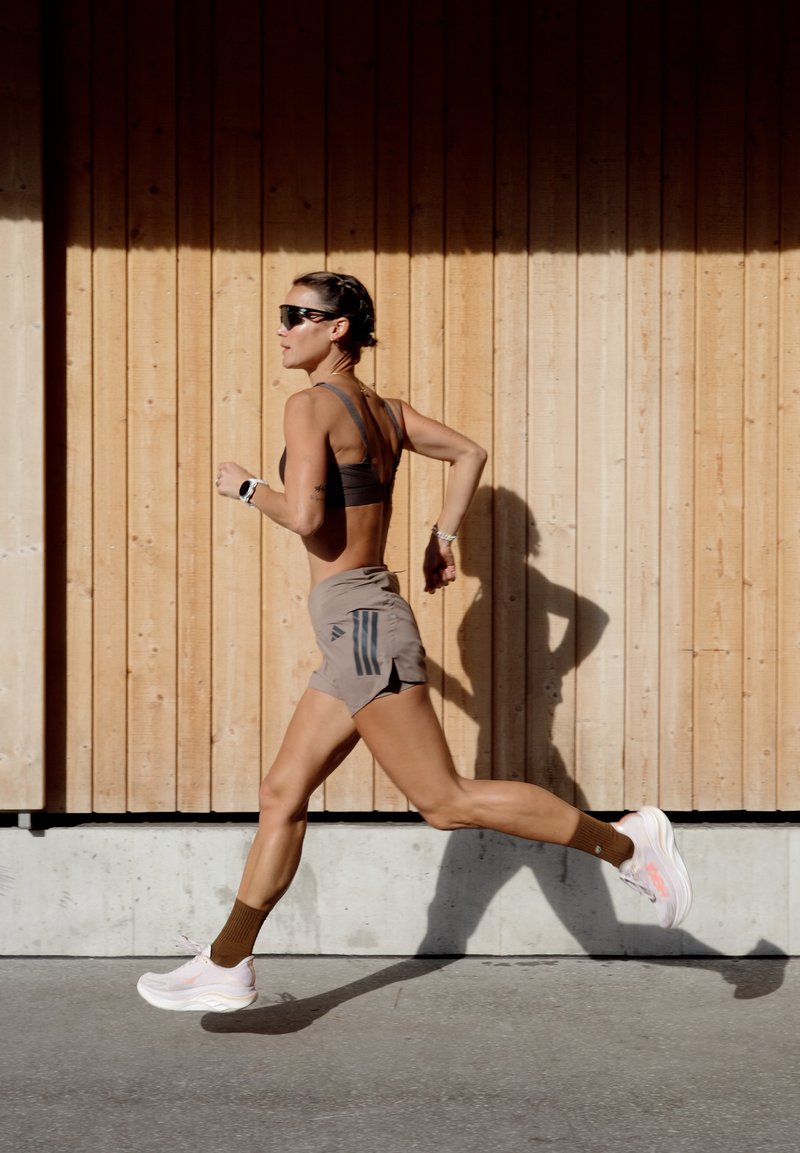 Femme en forme portant des lunettes de soleil et des vêtements de sport court rapidement devant un mur en bois en pleine lumière du jour, projetant une ombre au sol.