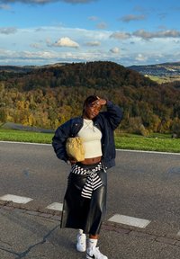 Casaco bomber preto, top curto bege, saia de látex preta com um laço listrado na cintura, ténis brancos, segurando uma mala bege, em pé ao ar livre.