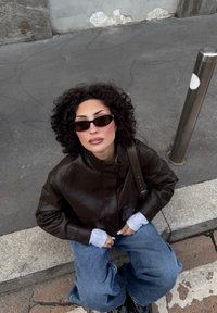 Brązowa skórzana kurtka, biała koszula z długim rękawem, za duże niebieskie dżinsy i ciemne okulary przeciwsłoneczne. Siedzi na chodniku w pobliżu metalowego słupa.