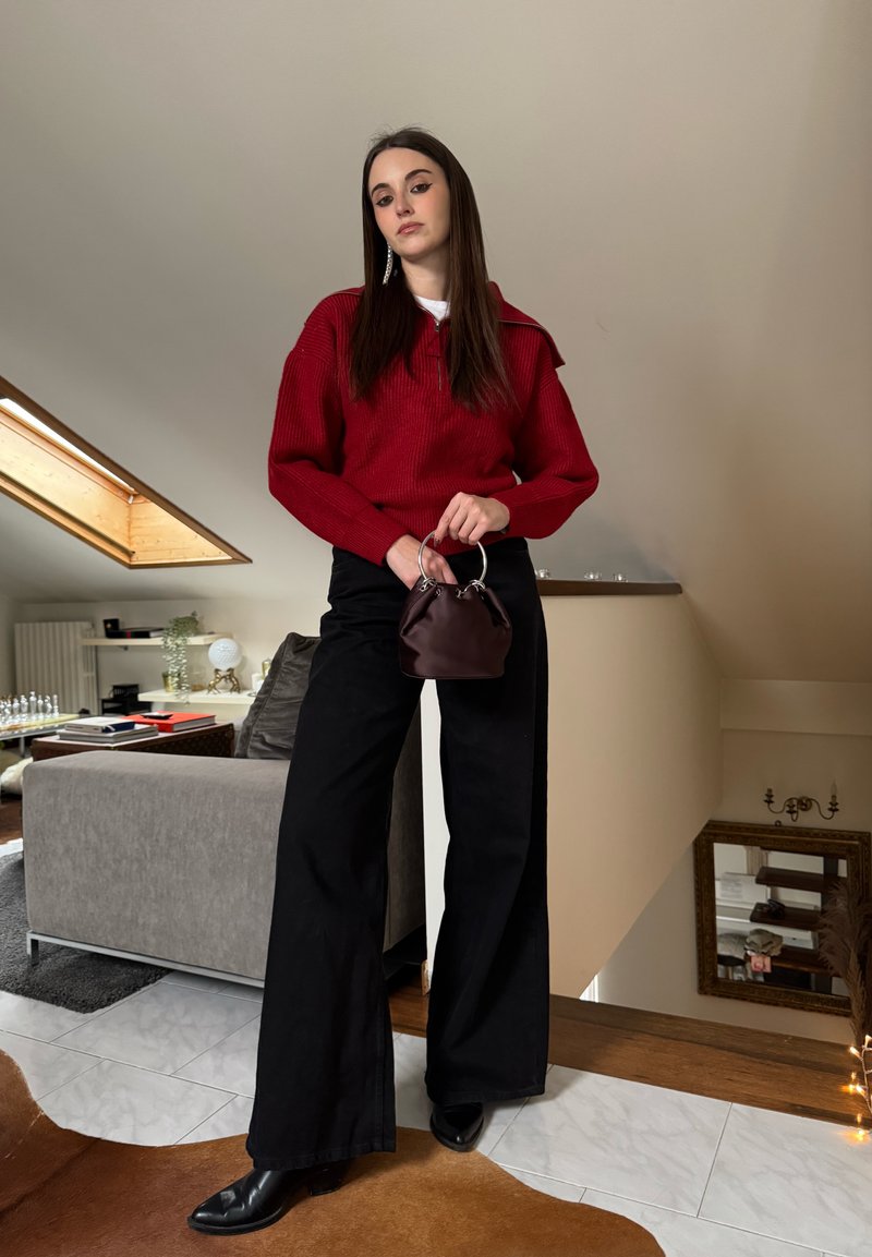 Jeune femme en pull rouge, pantalon noir à jambes larges et bottes noires, tenant un petit sac à main marron, debout dans un salon moderne.