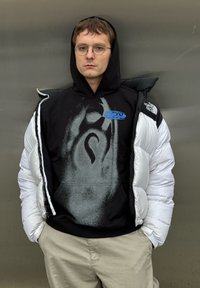 Zwarte hoodie met grafisch ontwerp, gedragen onder een witte pufferjas. Ronde bril, lichtgekleurde broek, staand tegen een grijze achtergrond.