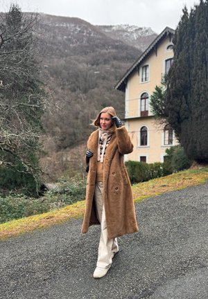 Mujer que lleva un abrigo largo marrón y guantes negros camina por una carretera cerca de una casa beige, con colinas boscosas y árboles al fondo.