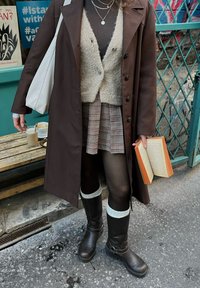 Brun langt yttertøy over en beige strikkegenser, rutete skjørt, sorte strømpebukser og brune støvler med hvite mansjetter til kneet, holder en bok.