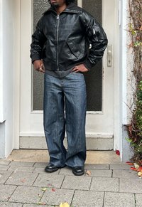 Melna ādas bumbvedēja jaka ar ribotiem aprocēm, kombinēta ar brīvām zilām džinsām. Melni apavi noslēdz tēlu uz neitrālas fona.