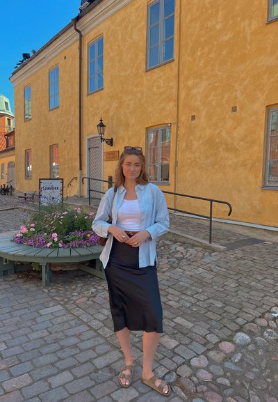 Camisa de botones azul claro sobre una camiseta blanca, falda de satén negra y sandalias marrones. Camino de adoquines con flores y edificios amarillos.