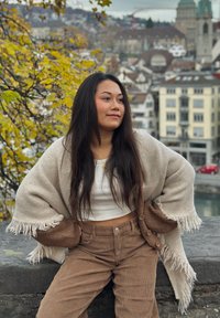 Châle en maille beige avec des bords frangés, pantalon en velours côtelé marron et haut blanc. Cheveux longs, assise contre un mur de pierre avec en arrière-plan la ville.