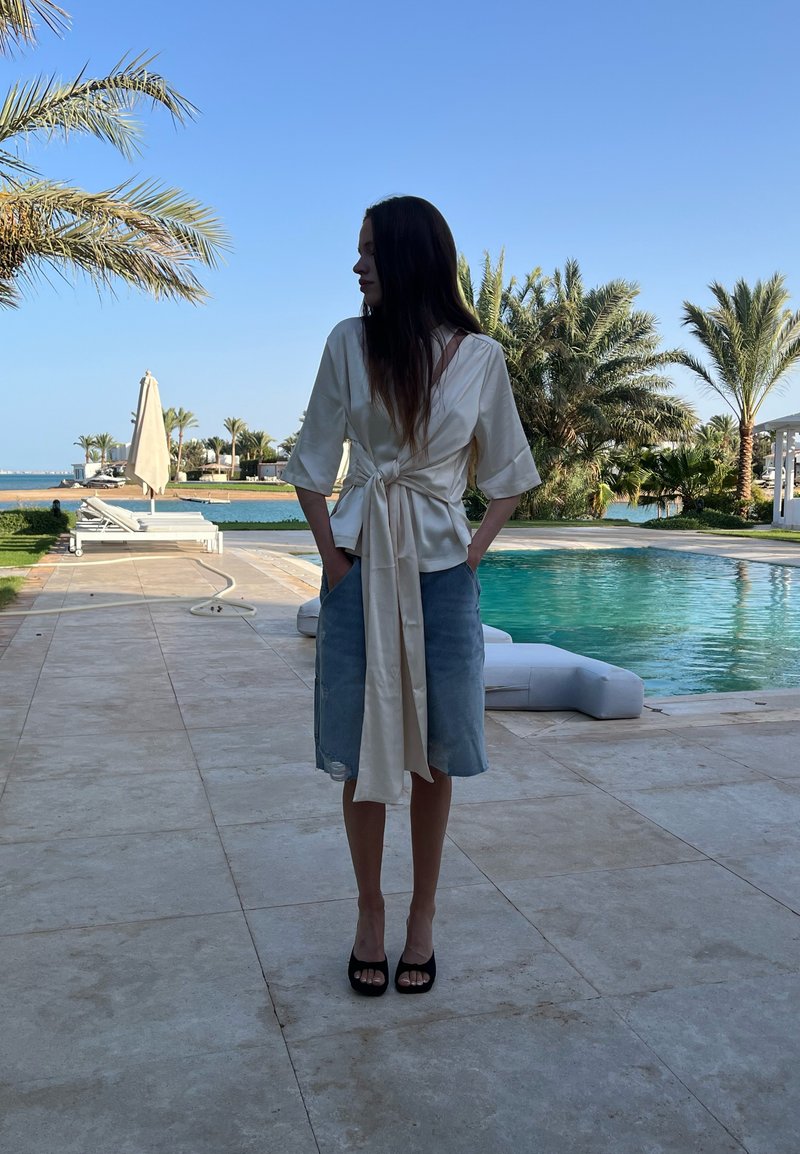 Κρεμ σατέν μπλούζα με φαρδιά μανίκια, δεμένη στη μέση, και μπλε τζην φούστα με φθαρτό τελείωμα. Μοντέλο που στέκεται δίπλα σε πισίνα περιτριγυρισμένη από φοίνικες.