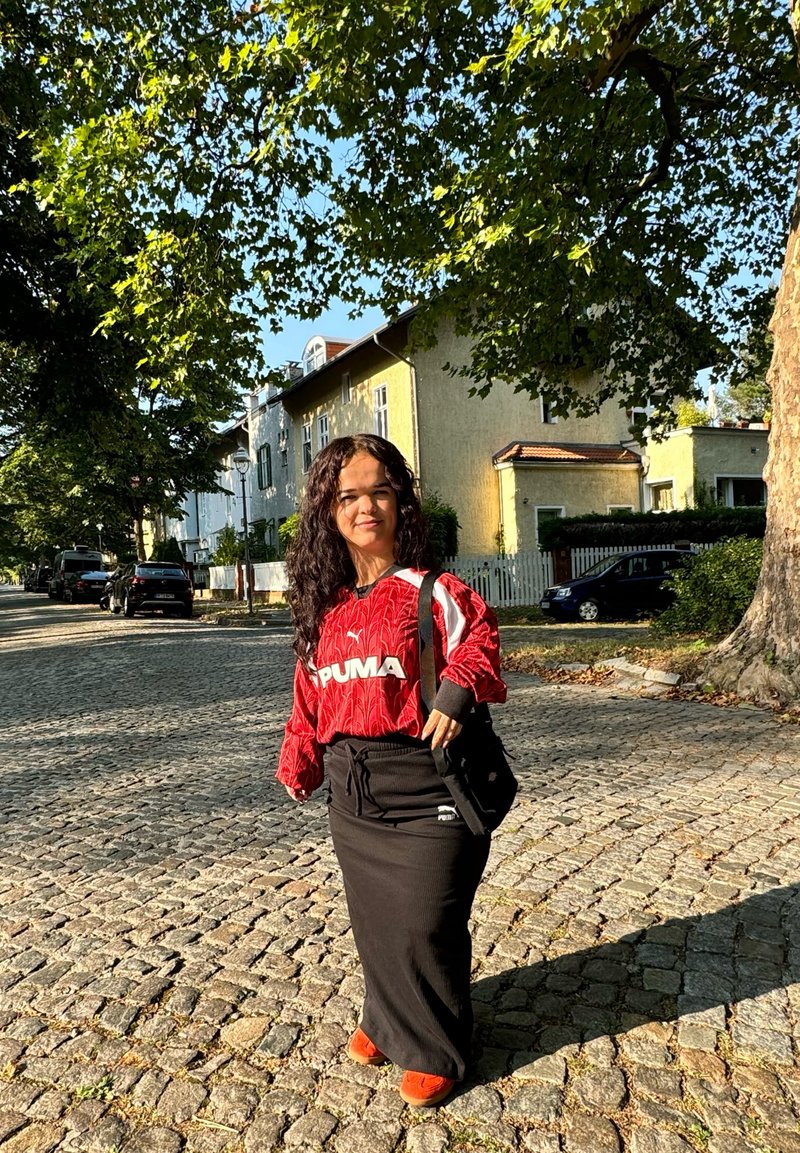 Κόκκινο μακρυμάνικο μπλούζα Puma με υφή, μαύρη μακριά φούστα και πορτοκαλί παπούτσια. Μαύρη τσάντα στον ώμο, στέκεται σε πεζόδρομο από cobblestones.