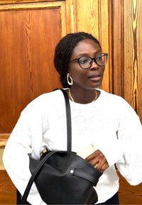 Kvinne med briller og gulløredobber som har på seg en hvit genser tar ut en lommebok fra en svart skulderveske foran en tre-dør.