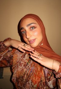 Hijab marrón, blusa con estampado floral en tonos cálidos, manos adornadas con múltiples anillos y joyas, maquillaje ligero y un ambiente interior.