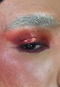 Nahaufnahme eines Auges mit kräftigem rotem und glitzerndem Lidschatten, silberner Augenbraue und glatter Hautstruktur, mit einem sanften rosa Akzent um das Auge.