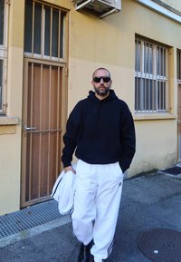 Vīrietis, ģērbjoties melnā kapuce, baltās Adidas sporta biksēs un saulesbrillēs, iet pa pilsētas trotuāru netālu no dzeltenas ēkas ar restotiem logiem un metāra durvīm.