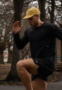 Čierne športové tričko s dlhým rukávom a krátke čierne šortky, �žltá čiapka, beh v parku s množstvom stromov v pozadí.