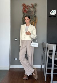 Korte witte blazer over een lichtgrijze top, gecombineerd met beige wijde broek en stevige bruine platform sandalen; mini witte handtas.