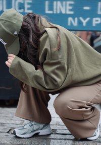 Sweat-shirt oversize vert olive, pantalon ample beige, baskets grises. La personne est accroupie, partiellement cachée, avec de longs cheveux visibles.