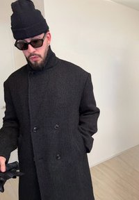 Homme portant un bonnet en maille noir, des lunettes de soleil foncées, un manteau noir texturé à double boutonnage, tenant des gants en cuir noir dans la main gauche.