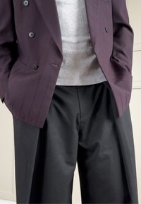 Doppelreihiger dunkelvioletter Blazer mit schwarzen Knöpfen, kombiniert mit einem grauen, gerippten Pullover und weiten schwarzen Hosen. Klare Linien, keine sichtbaren Muster.
