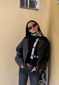 Mustast nahast jakk tõmblukkudega, must kõrge kaelusega sviiter, tumedad teksad ja mustvalge mustriga sall. Kandes tumedaid päikeseprille.