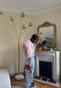Pink fluffy jacket, white top with a strawberry print, blue jeans, red handbag with charms, standing near a gold lamp and a mirror.