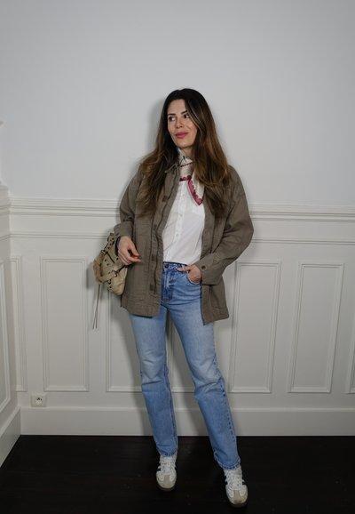 Chaqueta caqui sobre una camiseta blanca, jeans azul claros y zapatillas blancas. Los accesorios incluyen un bolso y un collar rosa. Fondo neutro.