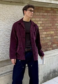 Burgundowa kurtka typu corduroy z kołnierzem i przednimi kieszeniami, połączona z czarną bluzką w prążki i ciemnoniebieskimi dżinsami o szerokich nogawkach. Trzyma książkę.