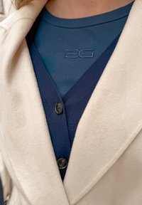 Άτομο που φοράει μπεζ παλτό πάνω από μια σκούρα μπλε πλεκτή ζακέτα με κουμπιά και μπλε ριγωτό τοπ με μικρό κεντημένο λογότυπο στο στήθος.