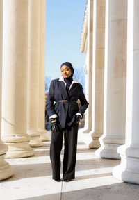Femme portant un hijab noir et un blazer à fines rayures avec ceinture, des gants noirs et un pantalon noir, debout entre des colonnes blanches en pierre classique à la lumière du jour.