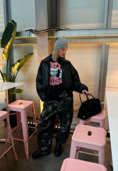 Cazadora bomber negra, sudadera gráfica negra y rosa, pantalones cargo de camuflaje y botas negras. Lleva una bolsa negra; gorro gris en la cabeza.