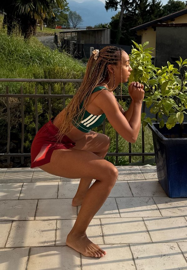 Une femme en tenue de sport maintient une flexion profonde des jambes sur une terrasse ensoleillée, à côté d’une plante en pot.