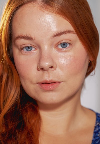 Retrato de una persona con cabello largo y liso de color rojo, ojos azules, piel suave y un aro en la nariz; vistiendo una blusa azul con un patrón. Iluminación suave.