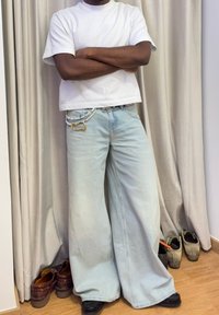 Lichtblauwe wijde jeans met een vervaagde afwerking, gedragen met een wit T-shirt. Een parelachtige kettingaccent is zichtbaar langs de tailleband.