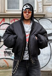 Doudoune noire avec fermeture éclair sur le devant et manches surdimensionn ées. Portée sur un t-shirt graphique, associée à un jean foncé et des chaînes argentées.