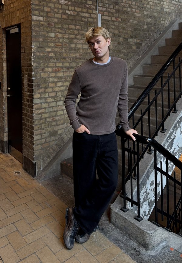 Mladý muž s blond vlasmi sa opiera o čierne kovové zábradlie schodiska v schodisku so steny z tehál, oblečený v hnedom svetri a čiernych nohaviciach.