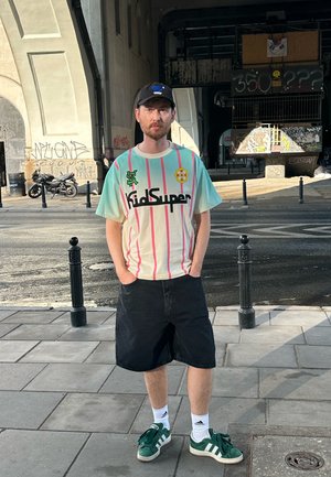 Camiseta gráfica de KidSuper con rayas verticales en rosa y menta, logo negro y acento amarillo. Shorts de mezclilla negros y zapatillas verdes de Adidas.