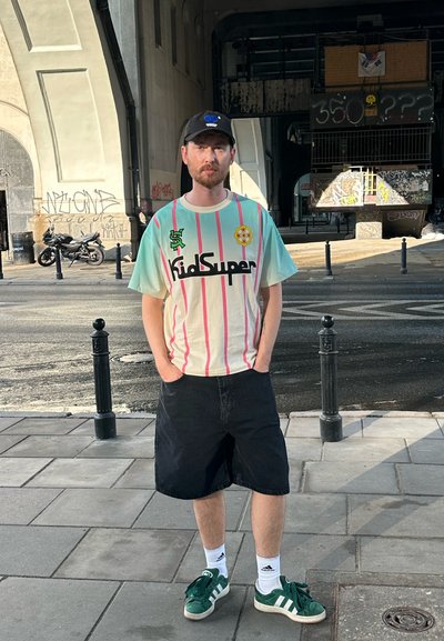 Camiseta gráfica de KidSuper con rayas verticales en rosa y menta, logo negro y acento amarillo. Shorts de mezclilla negros y zapatillas verdes de Adidas.