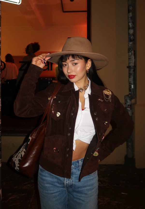 Jeune femme portant un chapeau marron, une veste marron avec des épingles, un crop top blanc noué et un jean bleu, posant à l'intérieur la nuit.