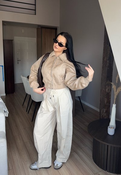 Chaqueta recortada de ante sintético beige con cremallera, cintura elástica y mangas oversized. Pantalones de pierna ancha color crema y zapatillas grises con detalles en blanco.