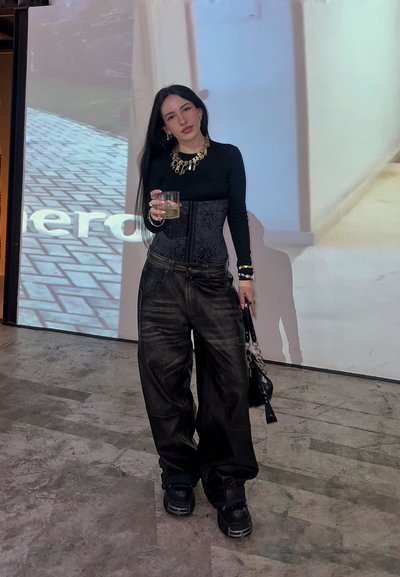 Top de corsé negro con detalles texturizados, pantalones oversized negros, zapatos gruesos negros, joyería en capas, sosteniendo una bebida, con un fondo claro.