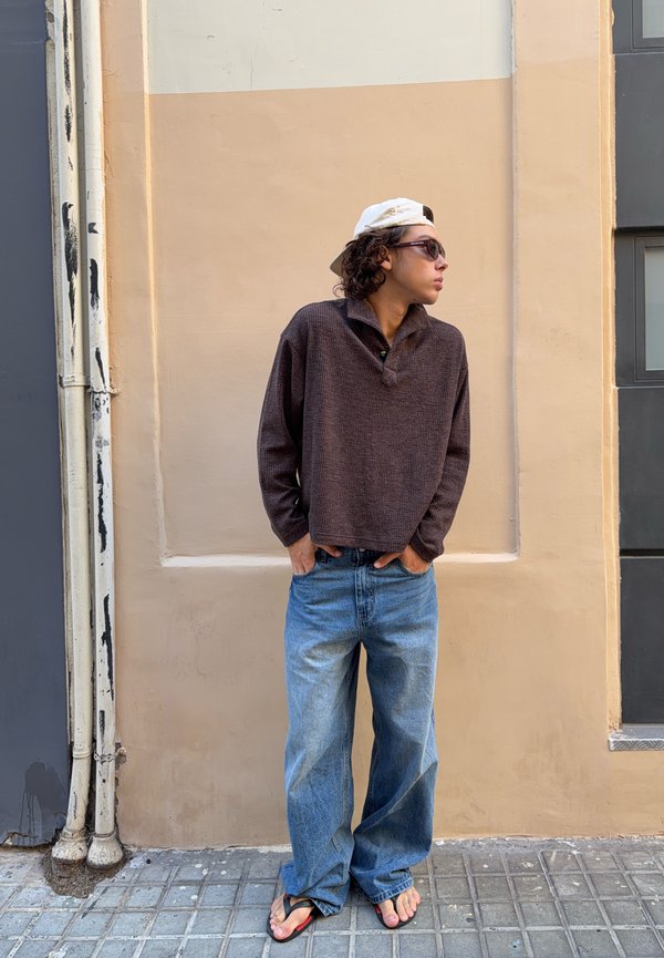 Ung person iført solbriller, hvid kasket bagvendt, brun sweater, vide jeans og flipflops står med hænderne i lommerne mod en beige væg.