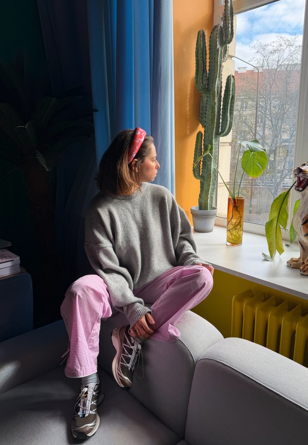 Femme portant un pull gris et un pantalon rose assise sur un canapé, regardant par la fenêtre avec une plante de cactus et un tigre en céramique sur le rebord de la fenêtre.