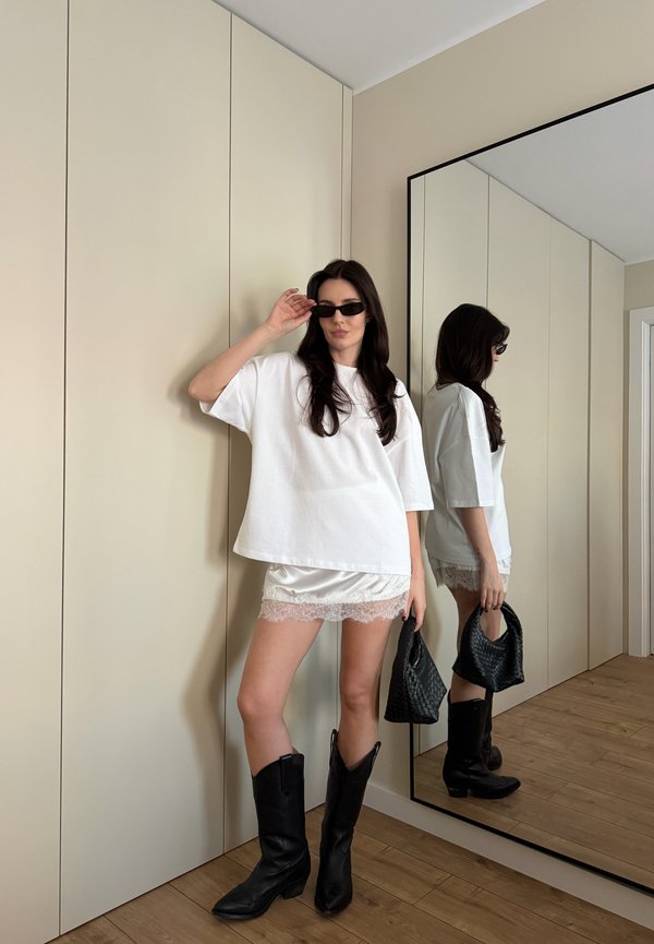 Mujer con camiseta blanca de gran tamaño, falda corta de encaje, gafas de sol negras, llevando bolso tejido negro y botas negras hasta la mitad de la pantorrilla, parada junto a un espejo.