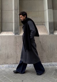 Jeune homme marchant le long d'un bâtiment en pierre, portant un long manteau sombre, un pantalon à jambes larges, une écharpe noire et des chaussures noires sur un trottoir carrelé.