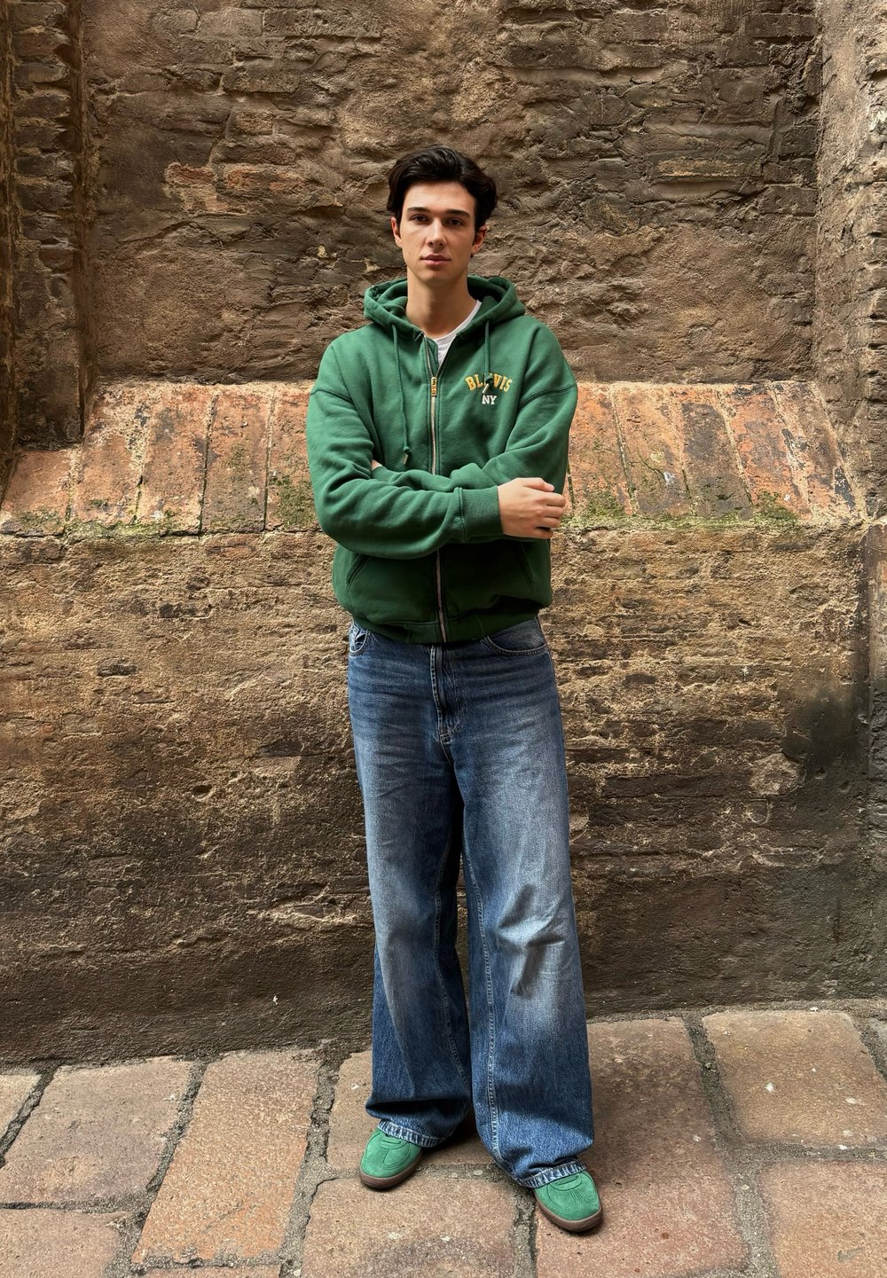 Green zip-up hoodie featuring yellow text, paired with loose blue jeans and green sneakers. Standing against a textured stone wall with arms crossed.